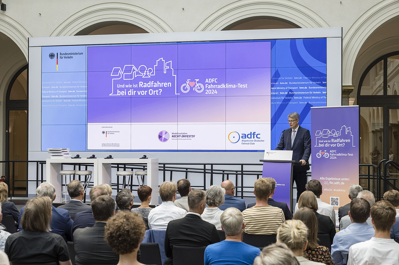 Ministervortrag Mann mit grauen Haaren und Brille in dunklem Anzug steht am Rednerpult vor großer lila Leinwand mit "Und wie ist Radfahren bei dir vor Ort? ADFC Fahrradklima-Test 2024", Publikum sitzt in Reihen davor