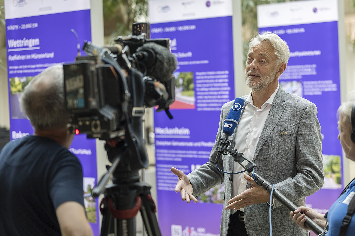 Medieninterview Frank Masurat, Mann mit grauen Haaren und Bart in hellgrauem Anzug, spricht in mehrere Mikrofone vor Fernsehkamera, im Hintergrund lila Banner mit Fahrradklima-Test-Informationen
