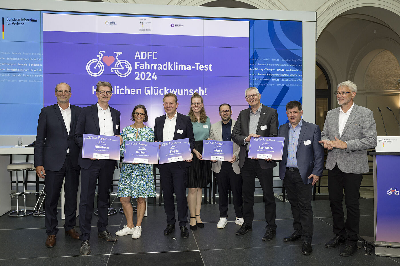 Preisverleihung Neun Personen stehen in einer Reihe vor großer Leinwand mit "ADFC Fahrradklima-Test 2024 Herzlichen Glückwunsch!" und halten lila Urkunden, darunter Nürnberg, Bochum, Lahr, Witten und Rheinbach