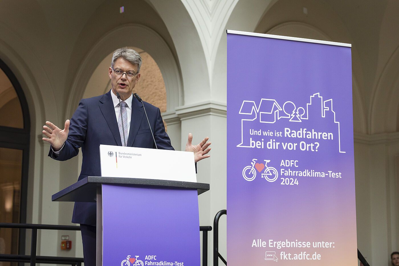 Minister am Rednerpult Mann mit grauen Haaren und Brille in dunklem Anzug gestikuliert am Rednerpult des Bundesverkehrsministeriums vor lila ADFC-Banner zum Fahrradklima-Test 2024
